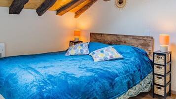 Ein Schlafzimmer mit schrägen Wänden, Holzbalken, einem Oberlicht, einem Bett mit blauer Bettwäsche, zwei Nachttischlampen und einem kleinen Nachttisch mit einer Lampe.