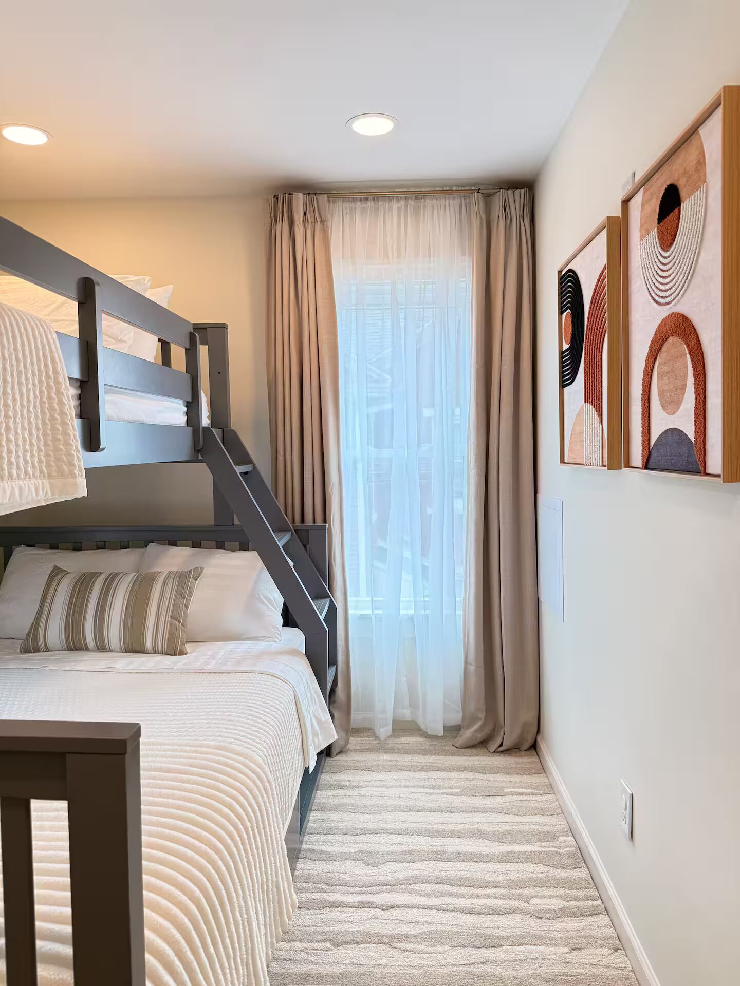 A bedroom with a bunk bed, a desk, a chair, a window with curtains, and abstract wall art.