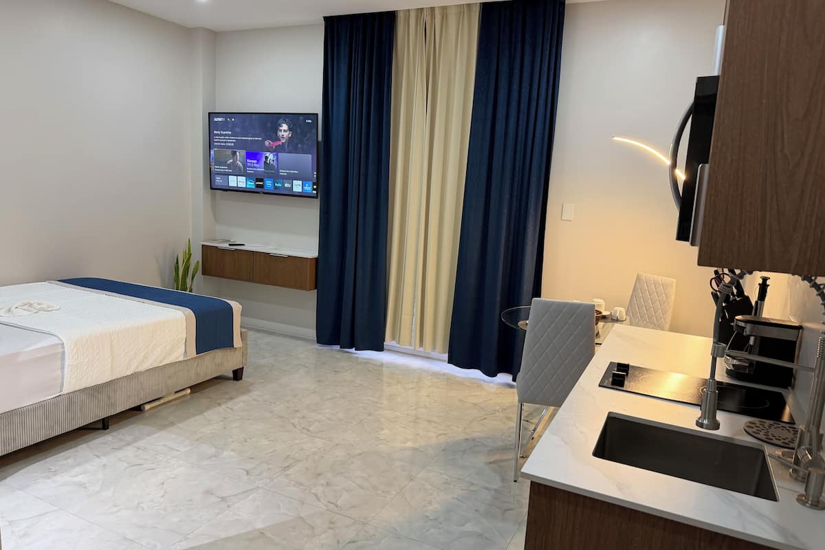 A hotel room with a bed, a desk with a sink, a chair, a TV mounted on the wall, and a window with curtains.