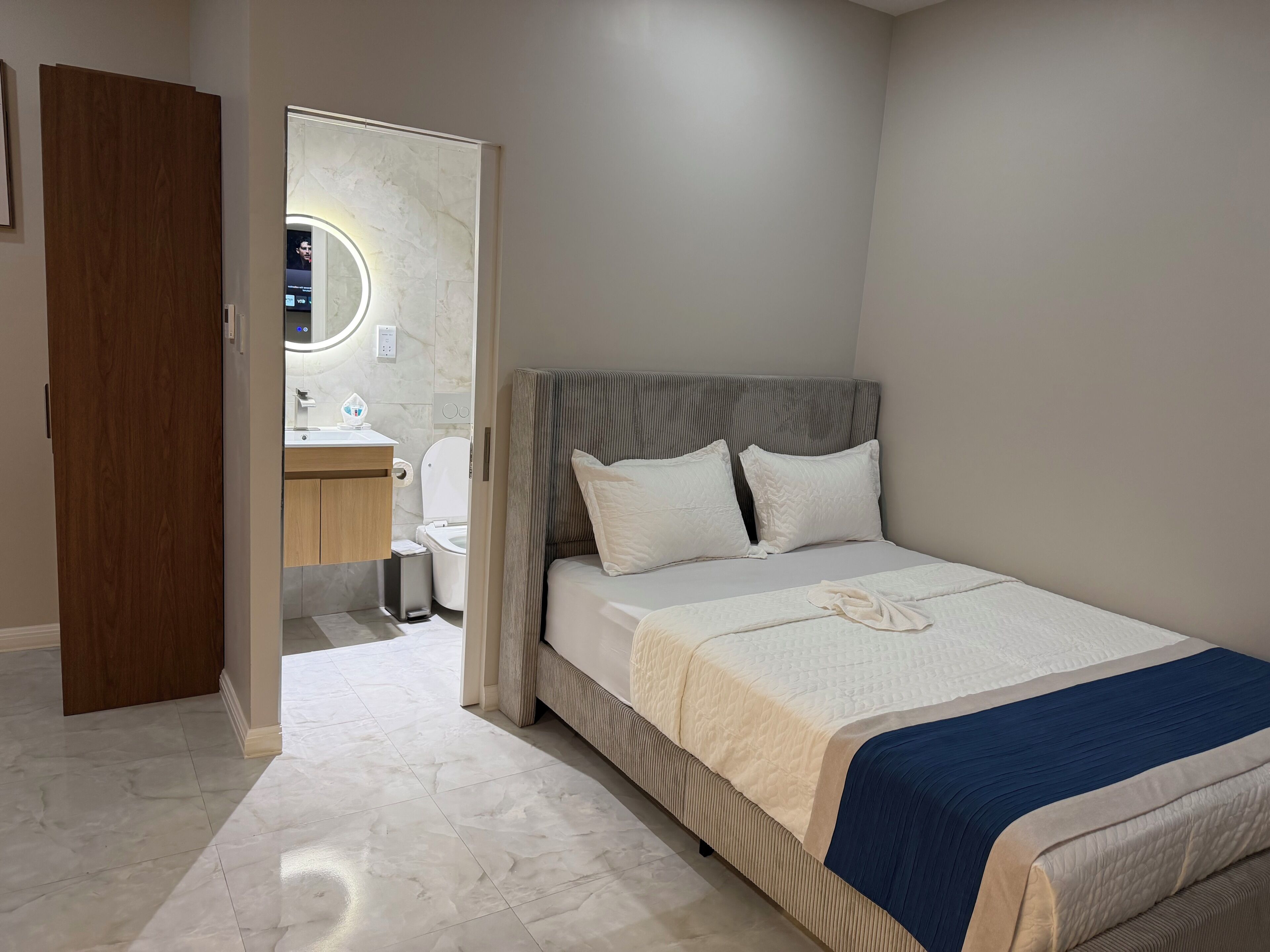 A bedroom with a bed, a bedside table, a mirror, and a bathroom visible through an open door.