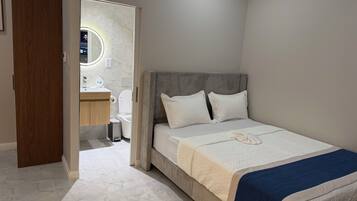 Un dormitorio con cama, mesita de noche, espejo y baño visible a través de una puerta abierta.