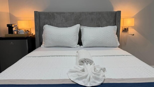 A neatly made bed with a folded towel, two pillows, and a headboard with a light fixture on either side. - Sanrai (Gros Islet)