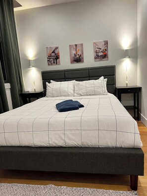 Ein ordentlich bezogenes Bett mit weißer und grauer Bettdecke, zwei Nachttischlampen und drei gerahmte Kunstwerke an der Wand.
