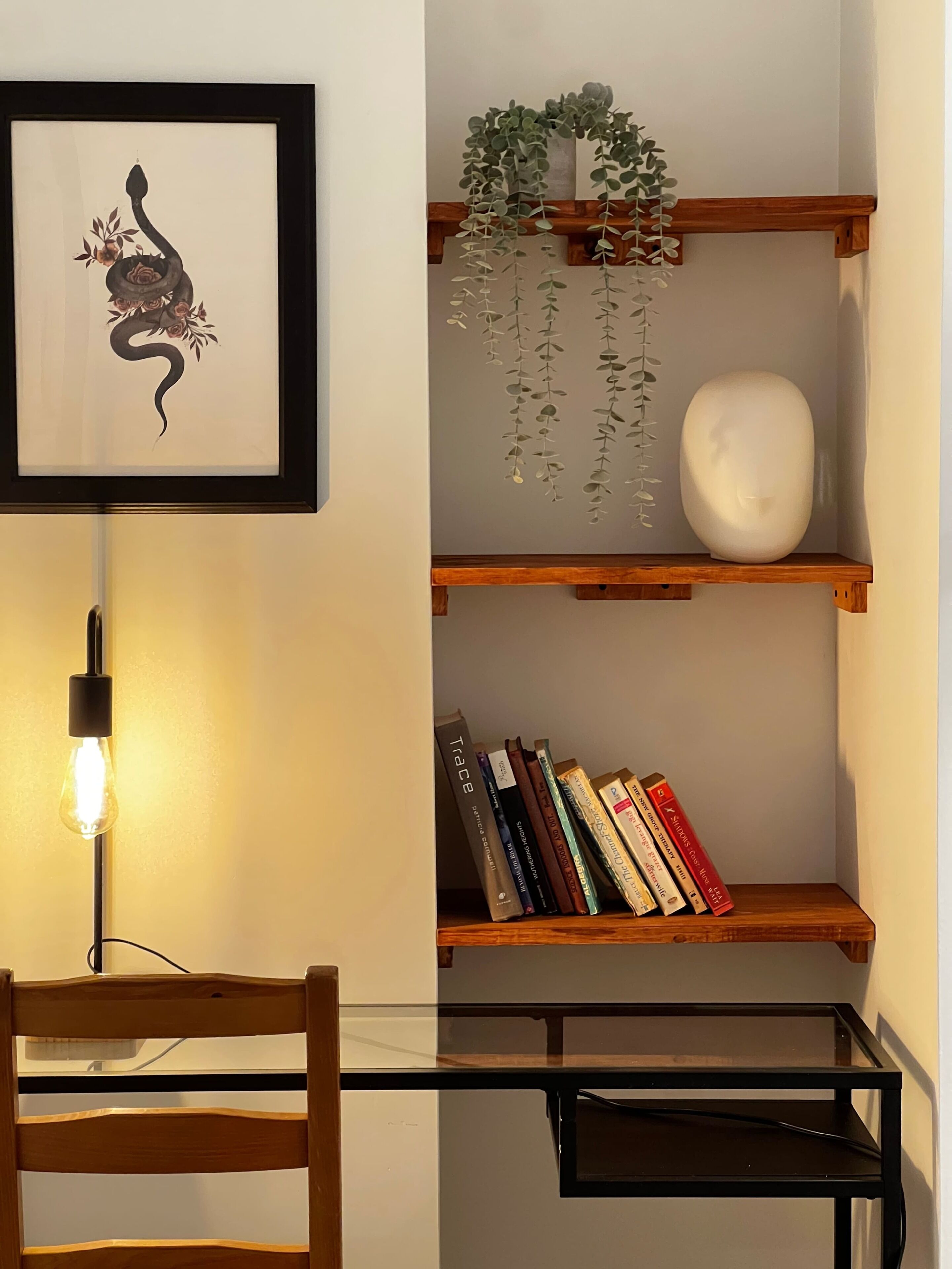 A room with a framed artwork, a hanging plant, a white vase, a bookshelf with books, and a glass table with a chair.