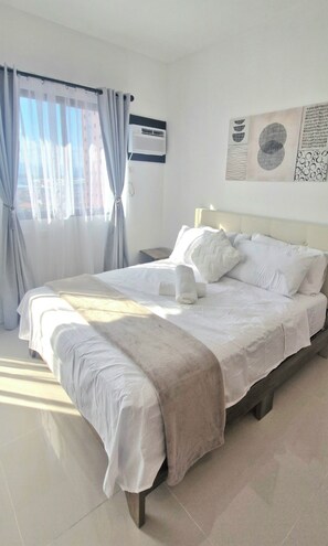 Een netjes opgemaakt bed met een witte deken en kussens, een nachtkastje met een lamp, een wandairconditioner en een raam met gordijnen.