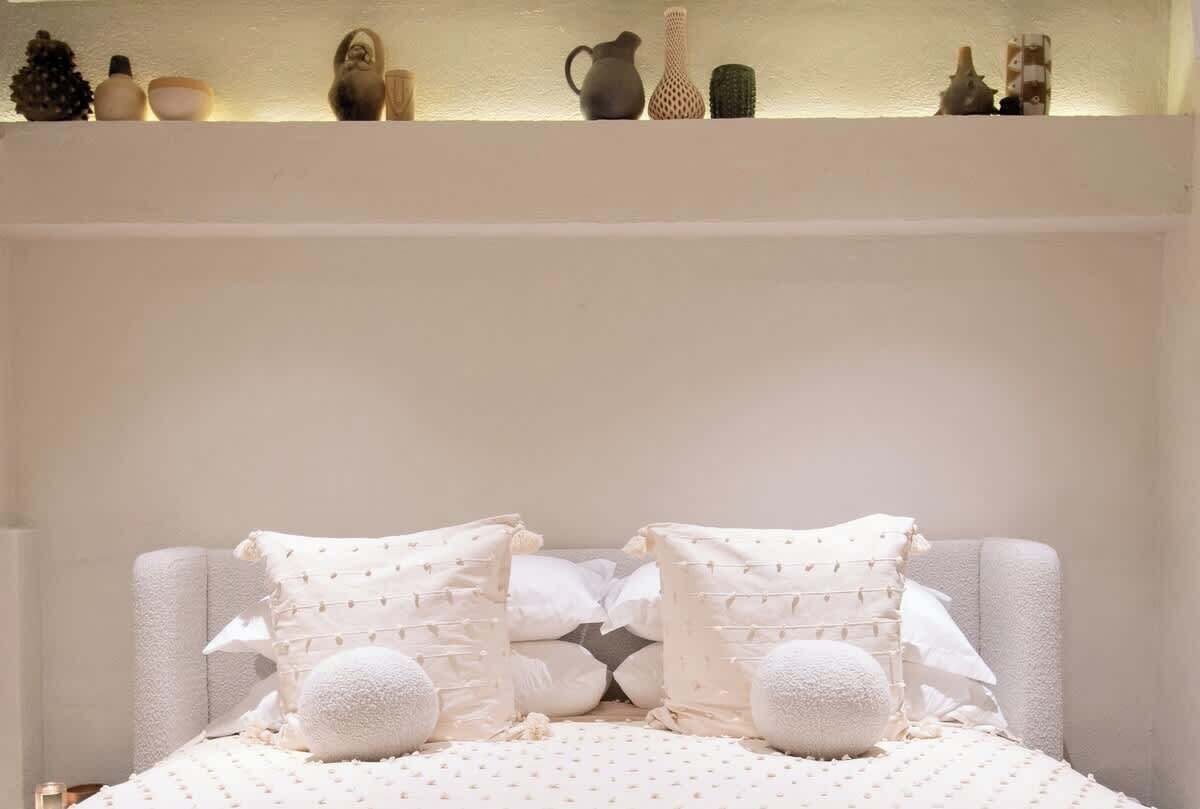 Un lit bien fait, recouvert d’une courtepointe blanche, agrémenté de deux coussins décoratifs, et une étagère au-dessus, où sont disposés divers objets.