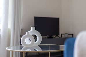 Ein modernes Zimmer mit einem Glastisch, zwei weißen abstrakten Skulpturen, einem Flachbildfernseher und einem blauen Sessel.