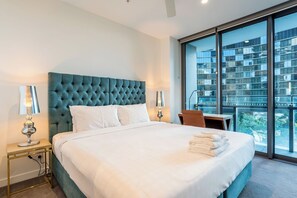 Ein Hotelzimmer mit einem großen Bett, zwei Nachttischlampen, einem kleinen Tisch und Blick auf ein Gebäude durch große Fenster.