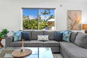 A grey sectional sofa with patterned cushions, a glass coffee table with a reed diffuser, and a view of palm trees through a large window.