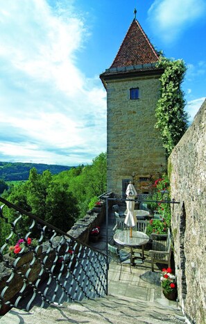 Una torre de piedra con techo de tejas, un ventanal pequeño y una escalera que lleva a una terraza con mobiliario exterior.