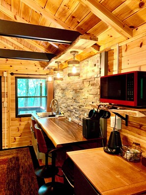 Interior de uma cabana de madeira com parede de pedra, teto de madeira, área de cozinha com micro-ondas, pia e mesa de jantar com cadeiras.