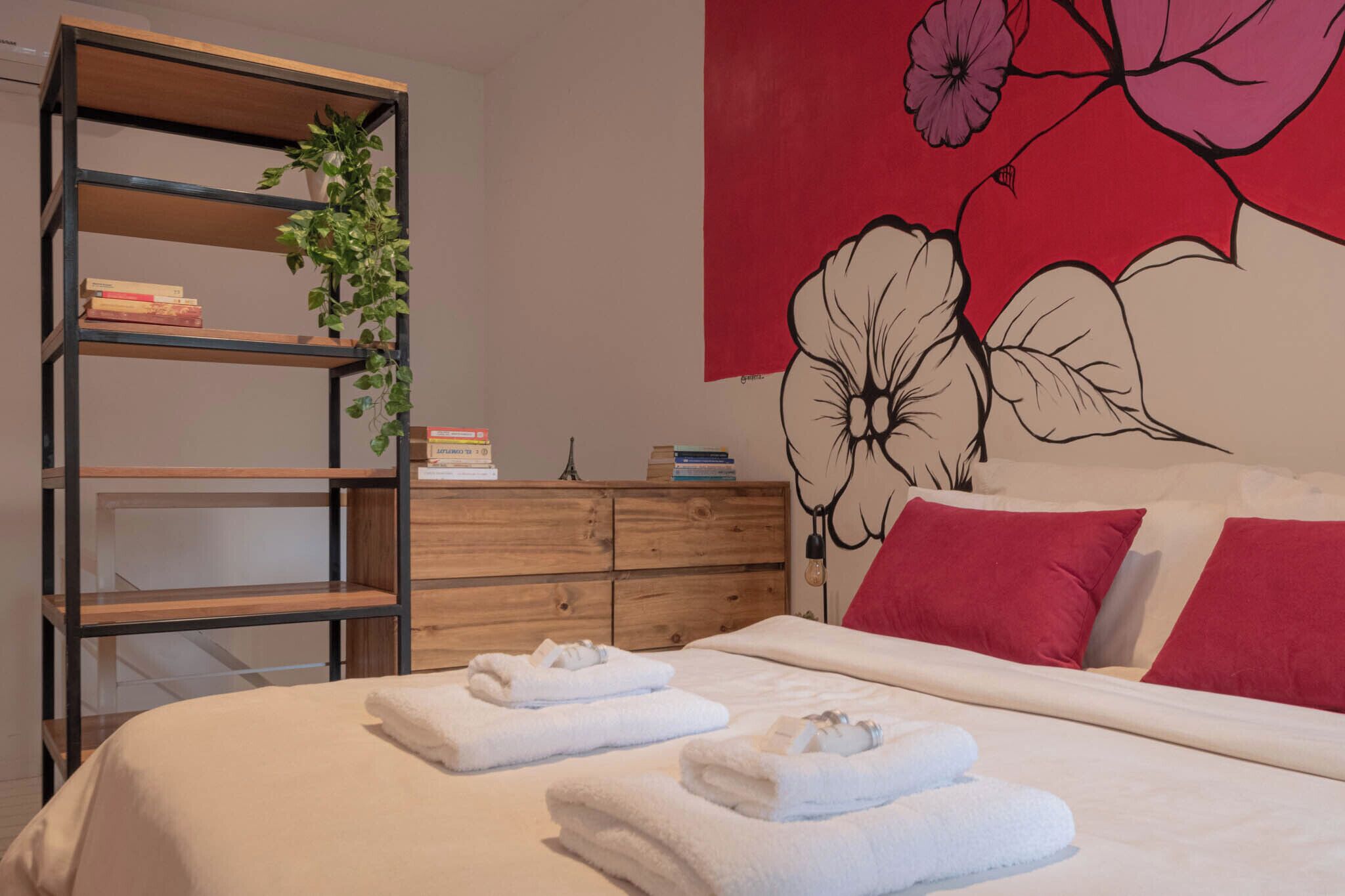Ein ordentlich bezogenes Bett mit weißen Leinen und Handtüchern, ein Holzregal mit Büchern, eine Holzkommode und eine Wand mit großem Blumenfries.