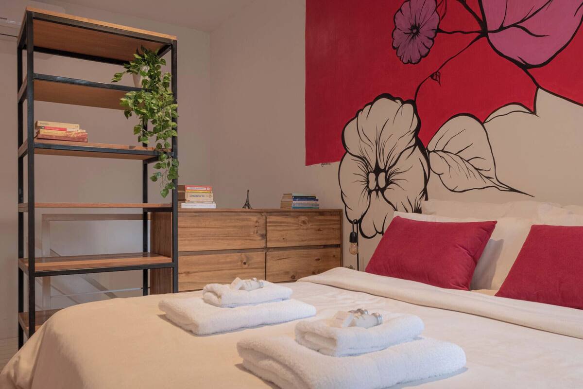 A neatly made bed with white linens and towels, a wooden bookshelf with books, a wooden dresser, and a wall with a large floral mural.