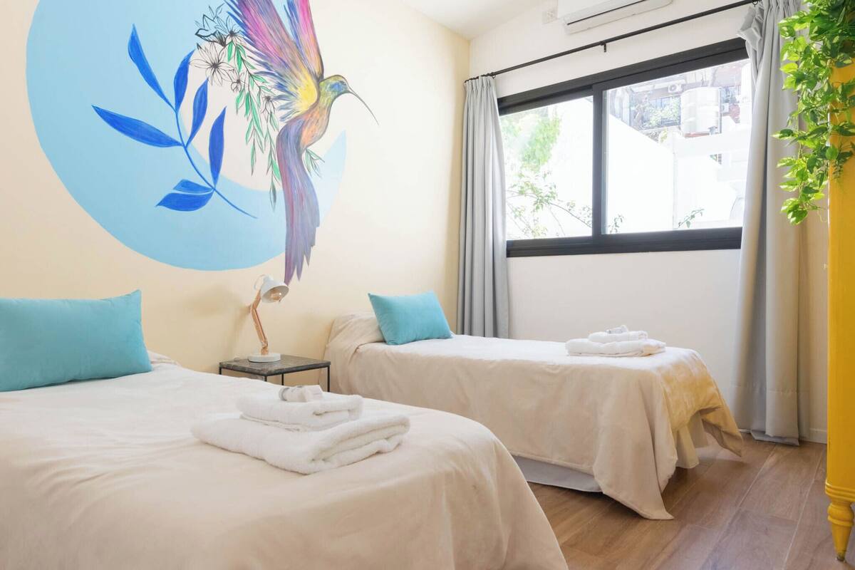 A hotel room with two beds, a window with curtains, a wall mural, and a plant.