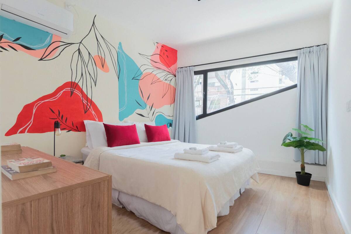 A modern hotel room with a large bed, a bedside table, a desk, a potted plant, and a window with curtains.