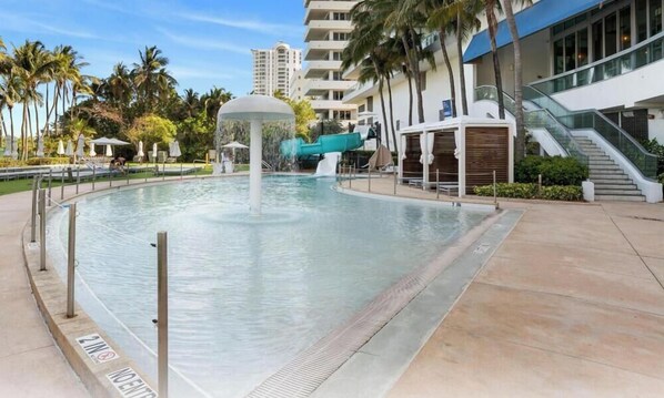 Uma piscina com uma fonte, cercada por palmeiras e um edifício moderno.