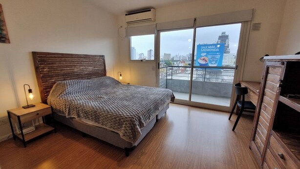 Een slaapkamer met een bed, nachtkastje, bureau en uitzicht op de stad door een groot raam.