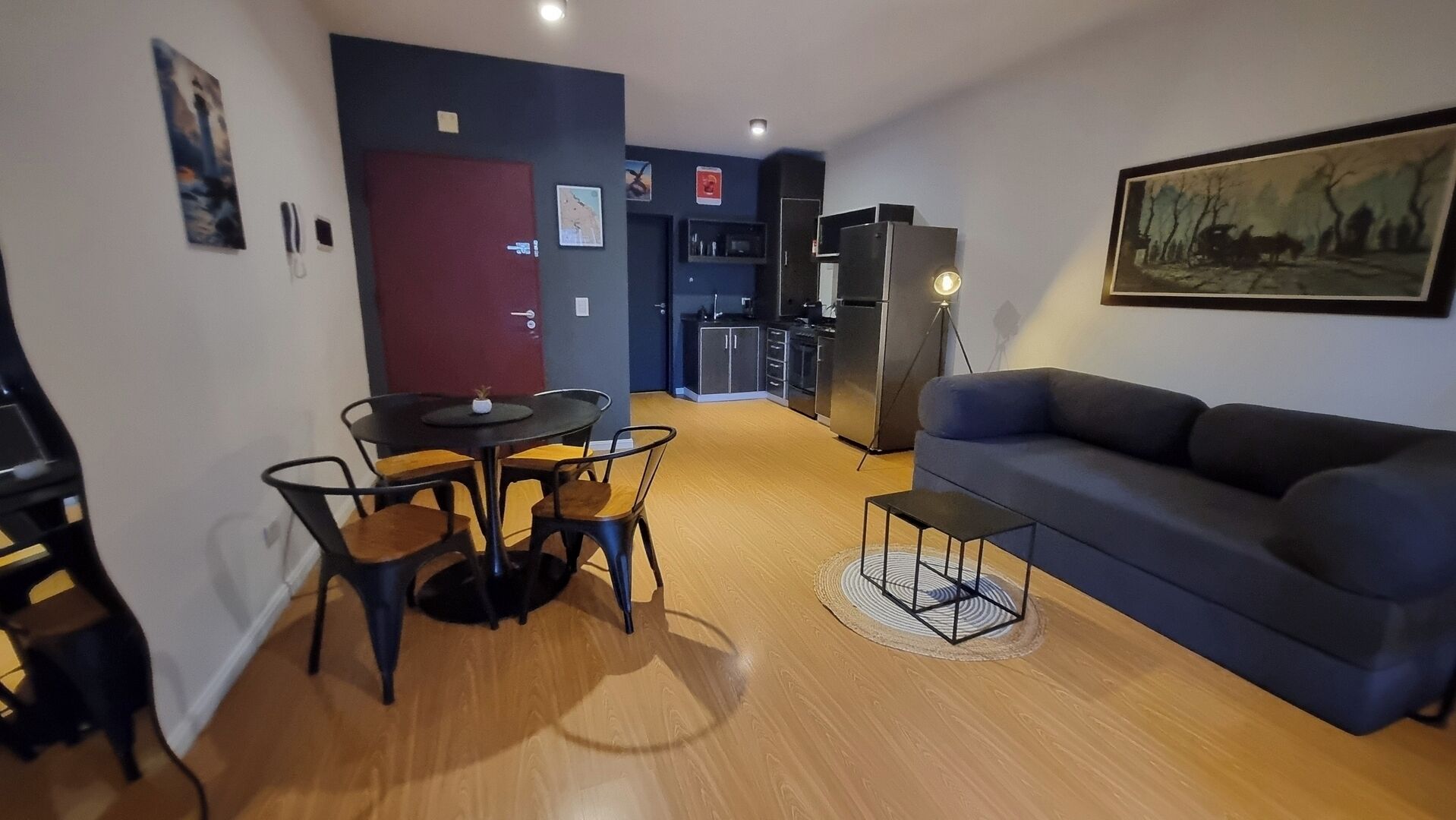 Una sala de estar moderna con un sofá, una mesa de comedor con sillas, una zona de cocina con electrodomésticos y un cuadro en la pared.