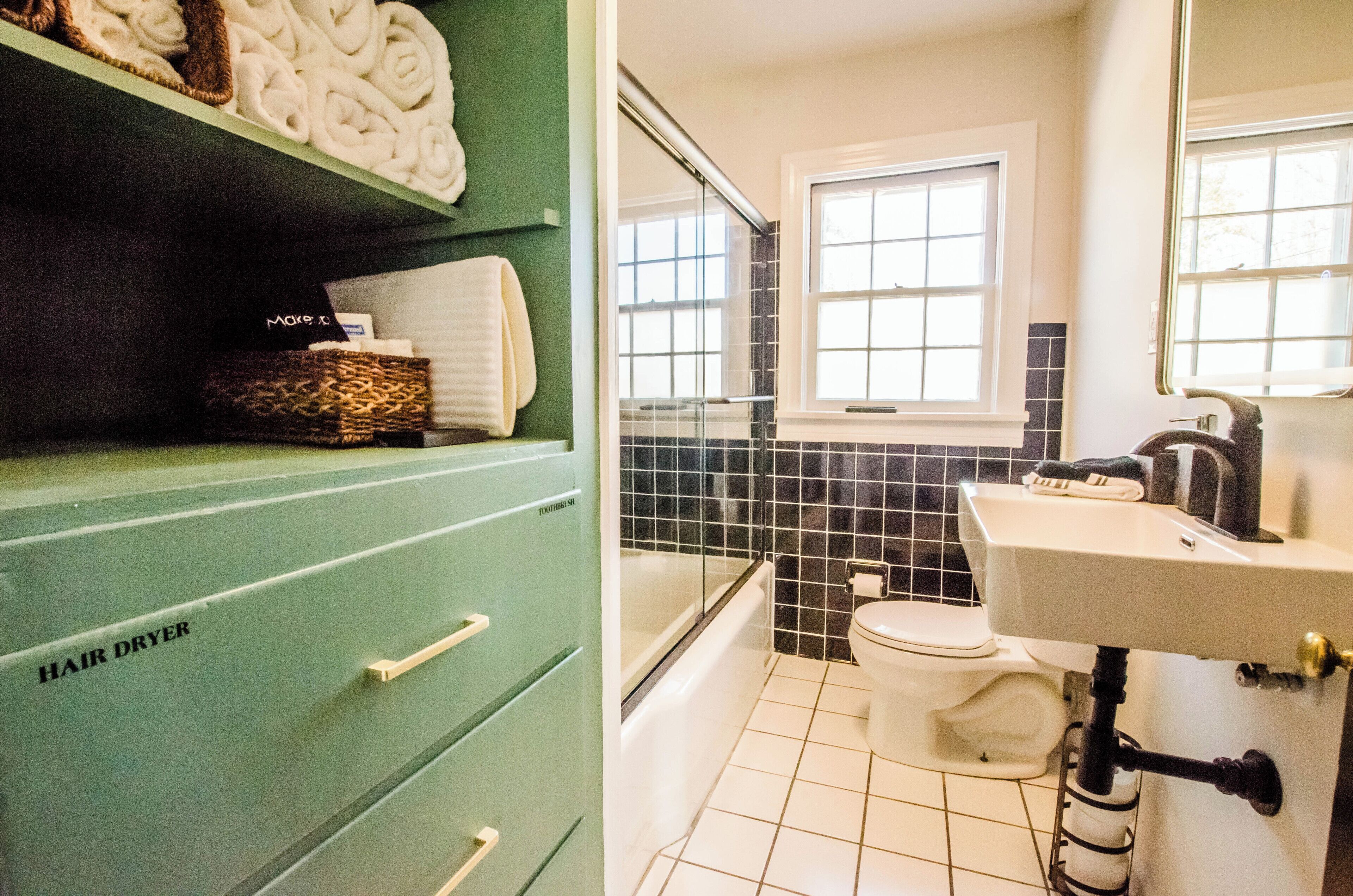 Une salle de bain avec un meuble de rangement vert sur lequel est inscrit « SÈCHE-CHEVEUX », un lavabo blanc, des toilettes et une fenêtre donnant sur l’extérieur.