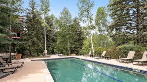 Una piscina con lettini e una terrazza, immersa tra alberi e un edificio.