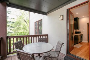 A covered patio with a round table and two chairs, a sliding glass door leading to a kitchen, and a view of trees outside. - Creekside Condo at the Base of Beaver Creek (Avon)