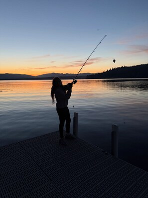 A person fishing at a dock with calm water and a sunset in the background.