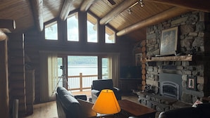 Interior acogedor de una cabaña con chimenea de piedra, un sofá de cuero, una mesa de madera con una lámpara y un ventanal con vistas a un lago.