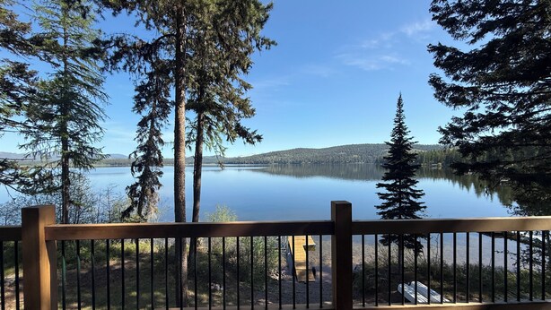 Una terraza de madera con barandilla da a un lago tranquilo, rodeado de árboles altos y montañas lejanas.