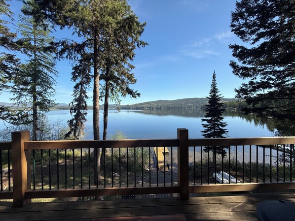 A wooden deck with a railing overlooking a calm lake, surrounded by tall trees and distant mountains.
