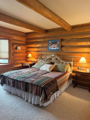 Un dormitorio de cabaña de troncos con una cama, dos mesitas de noche, un cuadro enmarcado en la pared y una ventana.