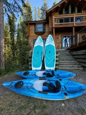 A wooden cabin with a deck, two paddleboards, and a blue kayak.