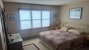 Un dormitorio con cama, mesita de noche, cómoda, ventana con persianas y un cuadro en la pared.