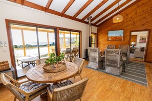 A wooden-floored room with wicker furniture, a round table with a plant centerpiece, and large windows offering a view. - Fourlees Breeze - Ocean Views - Sleeps 11 (Point Lookout)