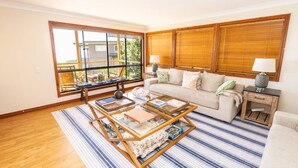 A living room with a sofa, a coffee table, and a window with blinds. - Fourlees Breeze - Ocean Views - Sleeps 11 (Point Lookout)