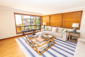 A living room with a sofa, a coffee table, and a window with blinds. - Fourlees Breeze - Ocean Views - Sleeps 11 (Point Lookout)