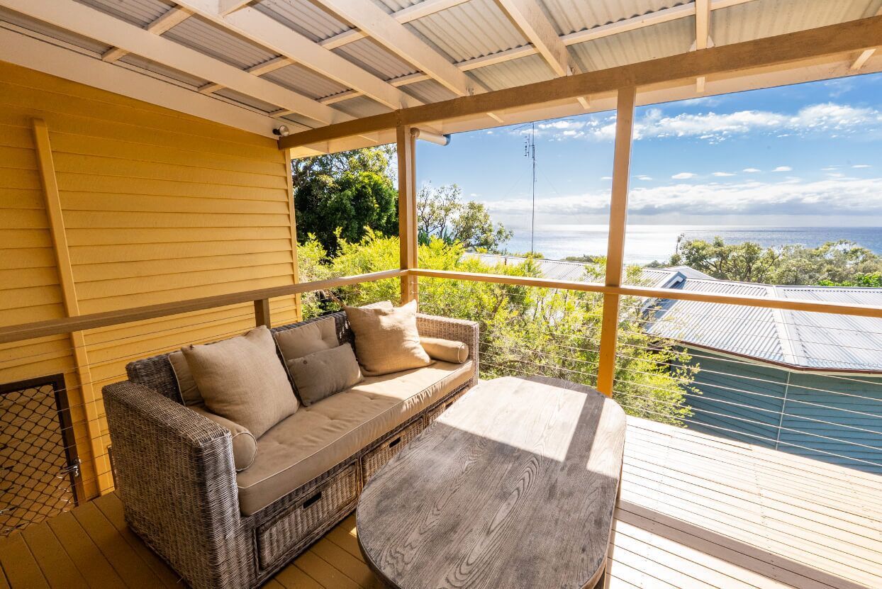 A covered outdoor seating area with a wicker sofa and a round table, overlooking a view of the sea and greenery.