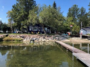 A lakeside cabin with a dock, surrounded by trees and a small rocky area.