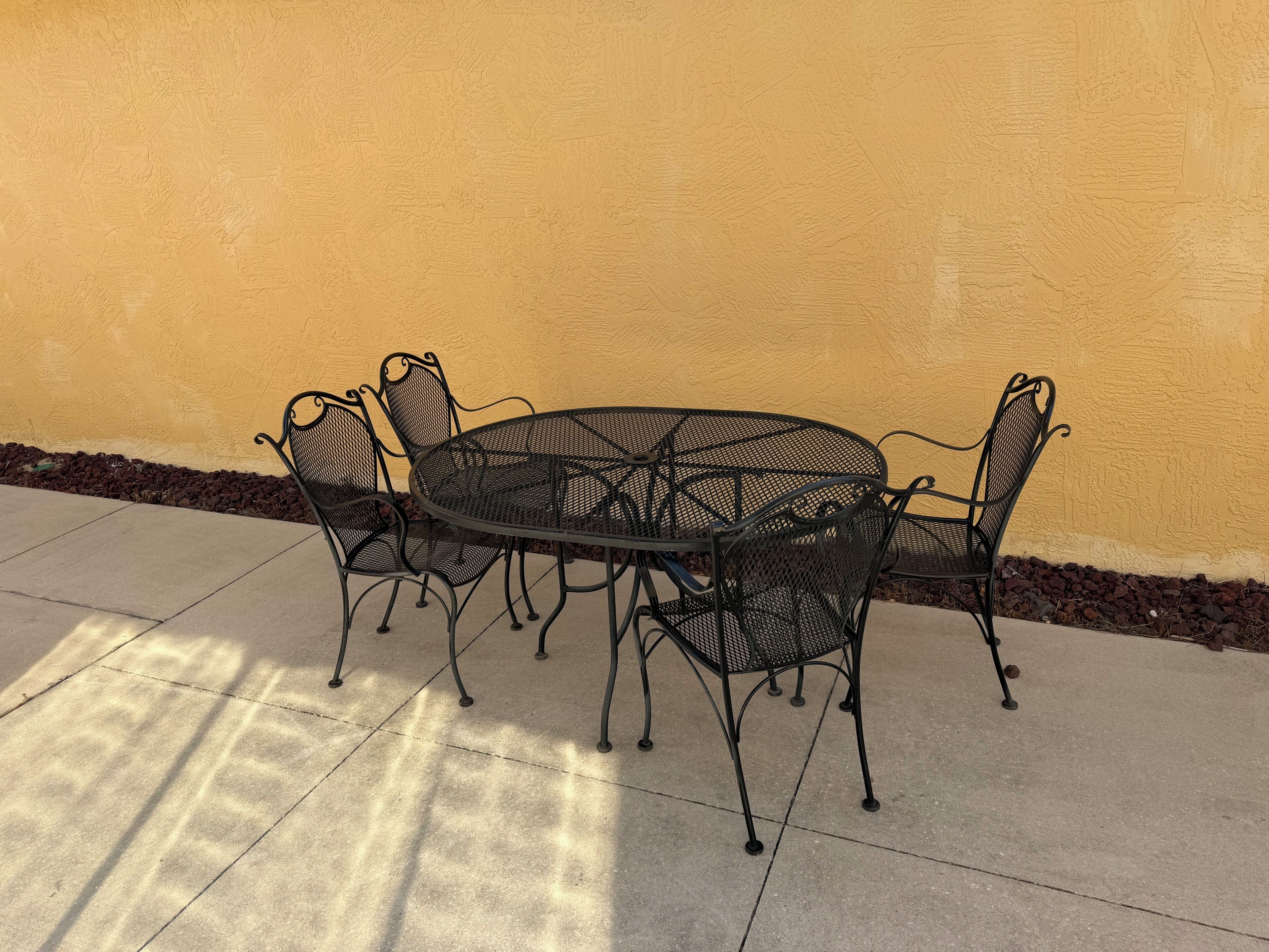 Ein runder Metalltisch mit vier Stühlen auf einer Beton-Terrasse vor einer gelben Wand.