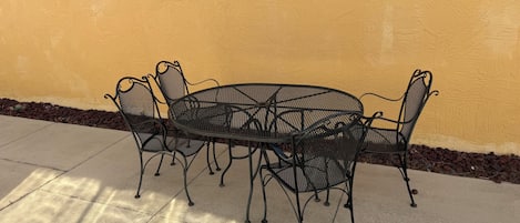 Ein runder Metalltisch mit vier Stühlen auf einer Beton-Terrasse vor einer gelben Wand.