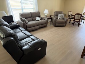 A living room with a sectional sofa, a recliner, a dining table with chairs, and a lamp on a side table. - Berea Villa's, Polo Ridge, Stunning Courtyard Villa, Golf Cart included! (The Villages)