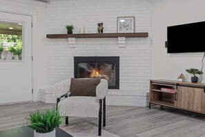 A living room with a white brick fireplace, a sofa, a TV mounted on the wall, and a wooden cabinet. - Cozy Luxury Getaway Home (Springfield)