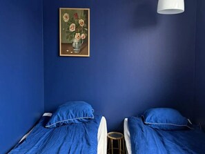 Ein Zimmer mit zwei Betten, einem kleinen Hocker, einem gerahmten Gemälde und einer weißen Lampe.