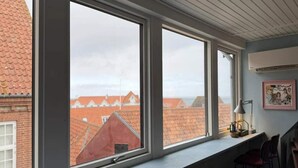 Una stanza con ampie finestre, vista su edifici con tetti rossi, una lampada e un quadro appeso al muro.