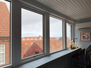 Ein Zimmer mit großen Fenstern, Blick auf Gebäude mit roten Dächern, einer Lampe und einem gerahmten Bild an der Wand.