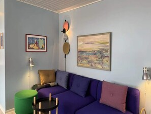 Ein Zimmer mit einem violetten Sofa, einem grünen Hocker, einem gerahmten Gemälde und einer Wandleuchte.