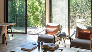 Ein modernes Wohnzimmer mit Couchtisch aus Holz, zwei Sessel, einem kleinen Beistelltisch und Blick auf einen Balkon mit Holzgeländer.