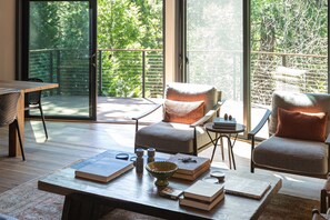 Una sala de estar moderna con una mesa de centro de madera, dos sillones, una mesita auxiliar y vistas a un balcón con barandilla de madera.