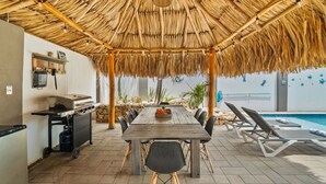 Un espace extérieur abrité, aménagé pour les repas, avec une table et des chaises, un barbecue, et une piscine en arrière-plan.