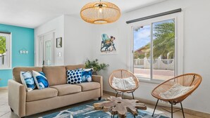 Un salon avec un canapé beige, deux fauteuils en osier, une table basse en bois, un tapis bleu à motifs étoilés, une grande fenêtre offrant une belle vue et un luminaire suspendu en osier.