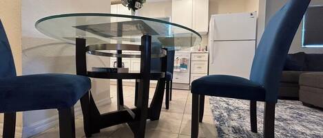 A dining area with a glass-top table, dark chairs, a vase with flowers, and a kitchen in the background.